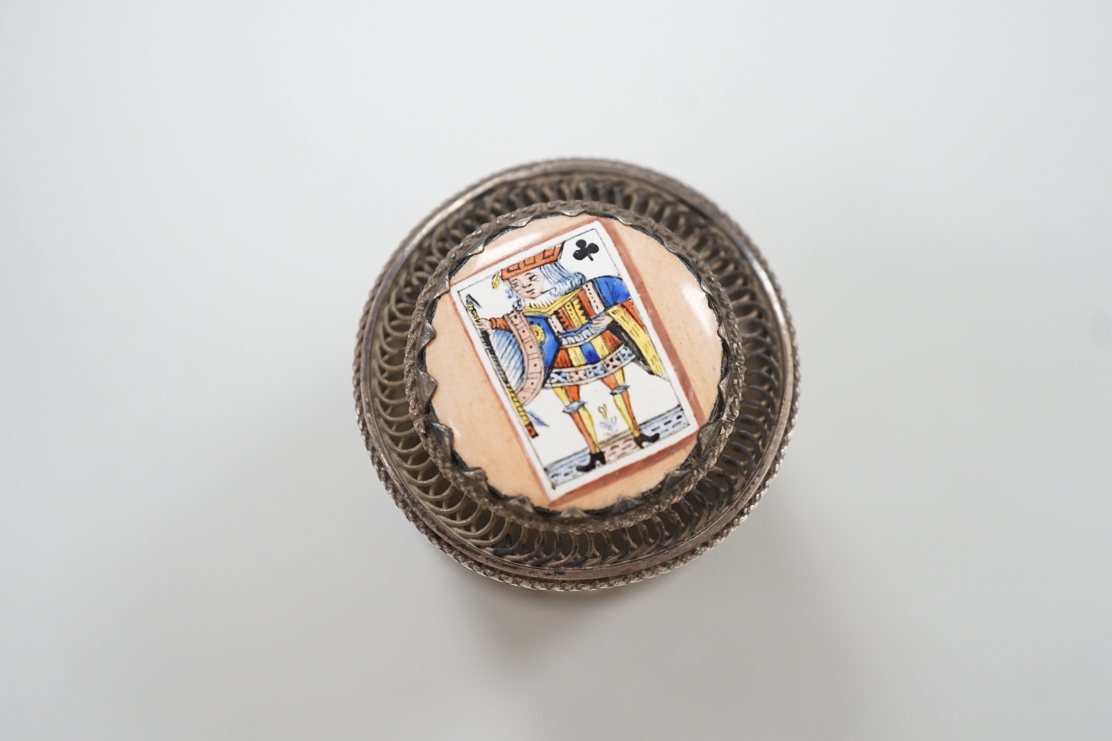 A continental filigree white metal and enamelled cylindrical pill box, the cover enamelled with a picture playing card, height 32mm.